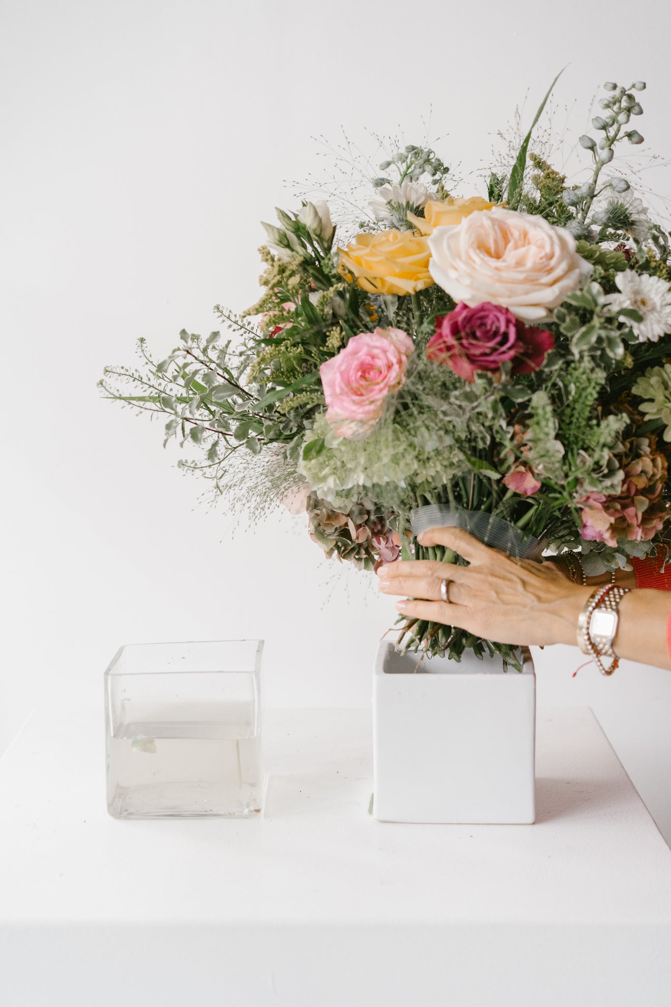 Bouquet of flowers held by a person over a white vase with an empty square vase of water next to it.